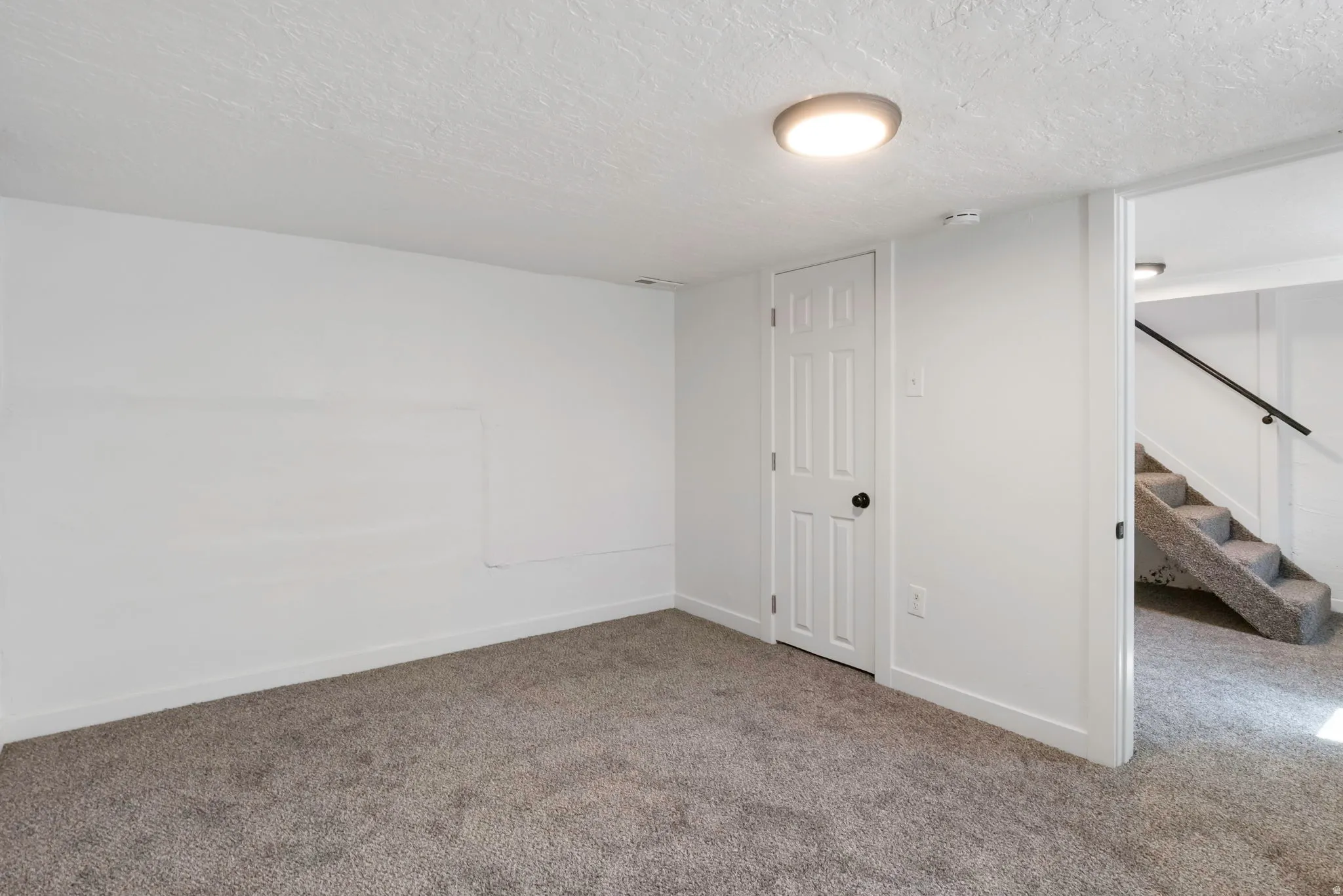 Carpeted room featuring a white paneled door with a black handle, a flush-mount ceiling light, and access to a carpeted staircase with a black handrail