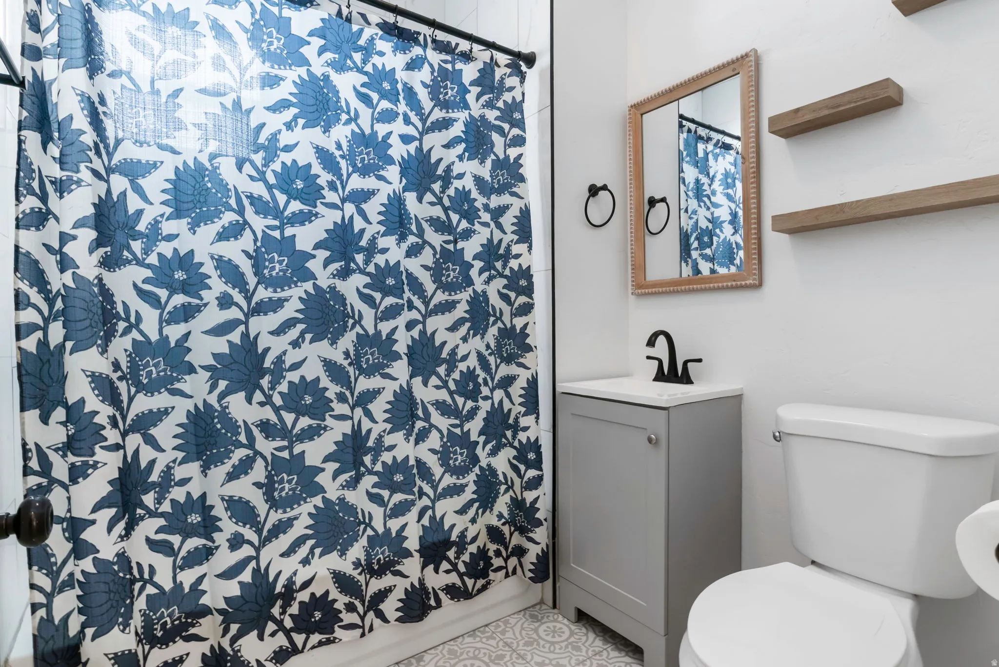 Bathroom featuring a vanity with a light gray cabinet and white countertop, black matte faucet, a framed mirror, and two wooden floating shelves