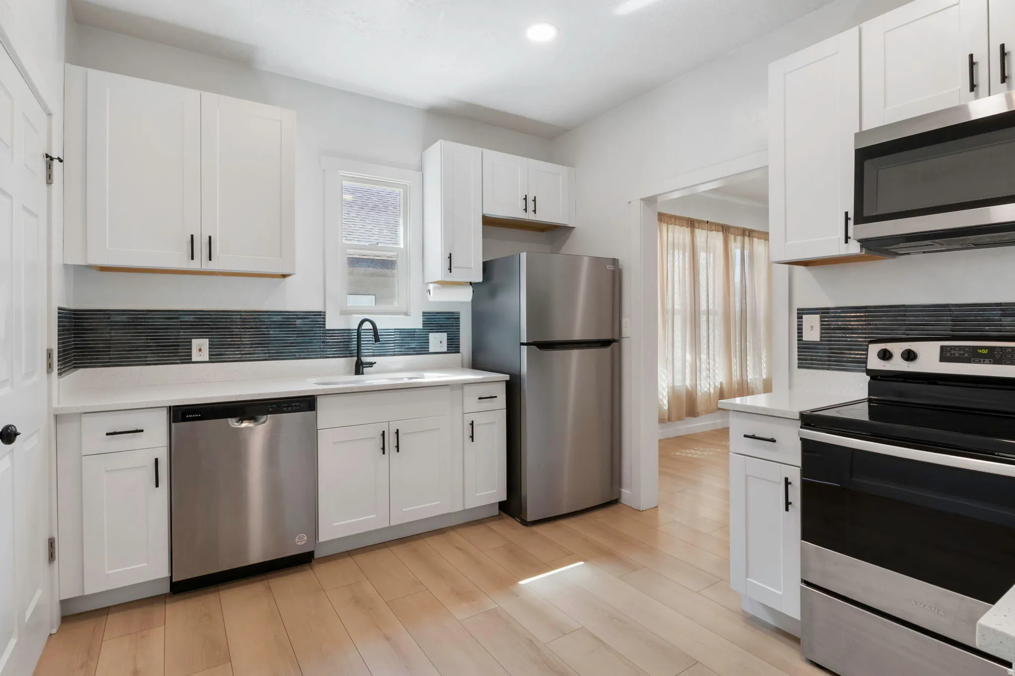Modern kitchen featuring light wood-finish flooring, white shaker cabinetry with matte black hardware, stainless steel appliances, a dark linear tile backsplash, and white countertops