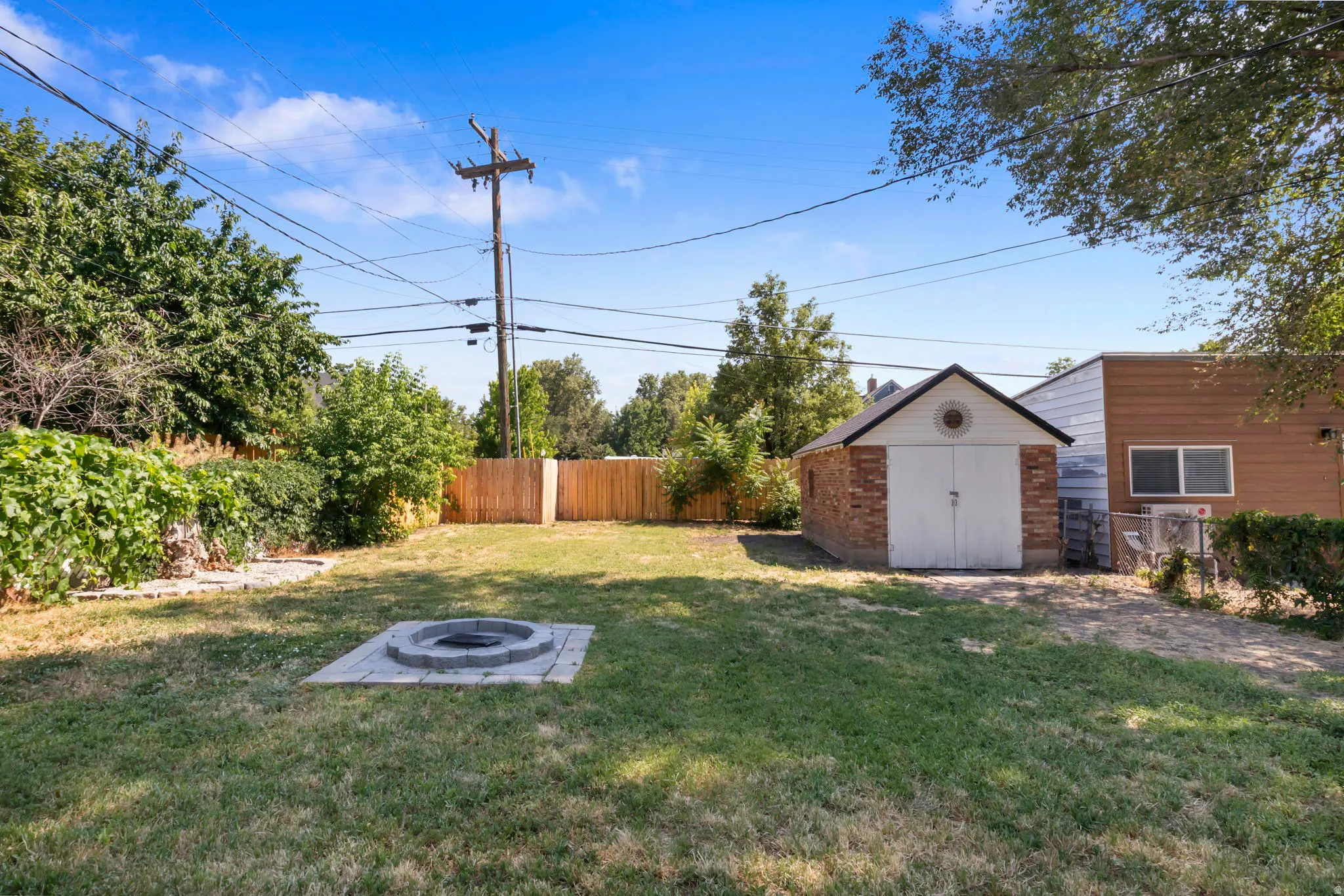 Expansive grassy backyard featuring a built-in fire pit with stone surround, a detached brick garage with white doors, wood fencing, and mature perimeter trees