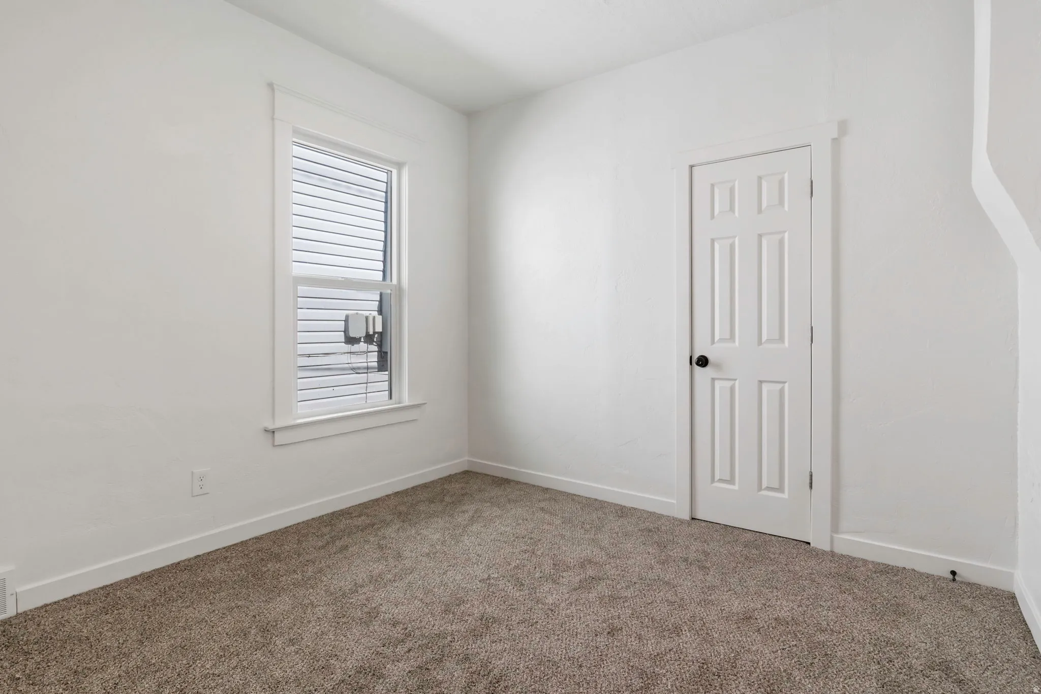 Room featuring a window with white blinds, a six-panel door with a black knob, light-colored walls, and textured carpeting