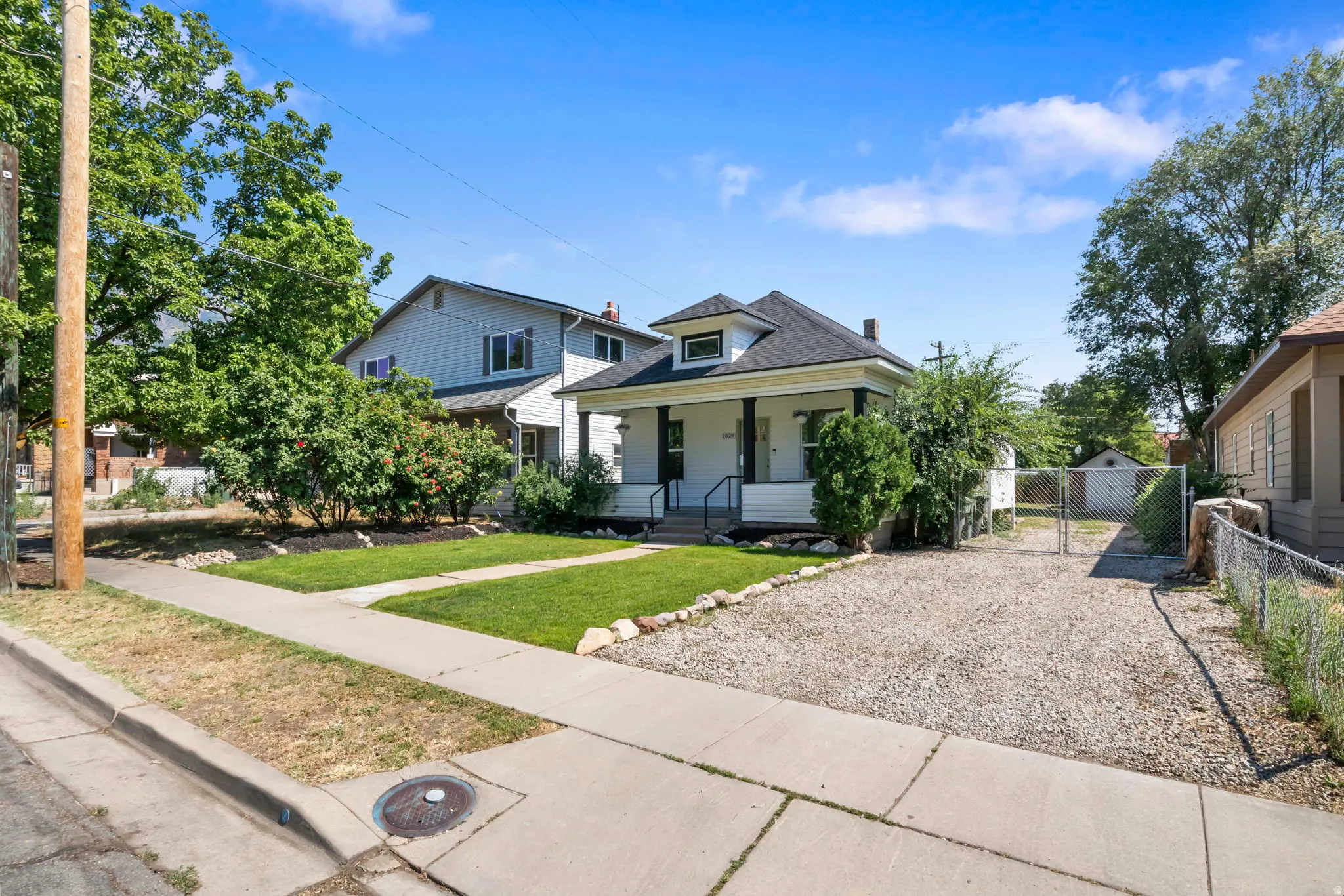 Classic white residence featuring a covered front porch with dark pillars, a gravel driveway, a grassy front yard, and mature landscaping
