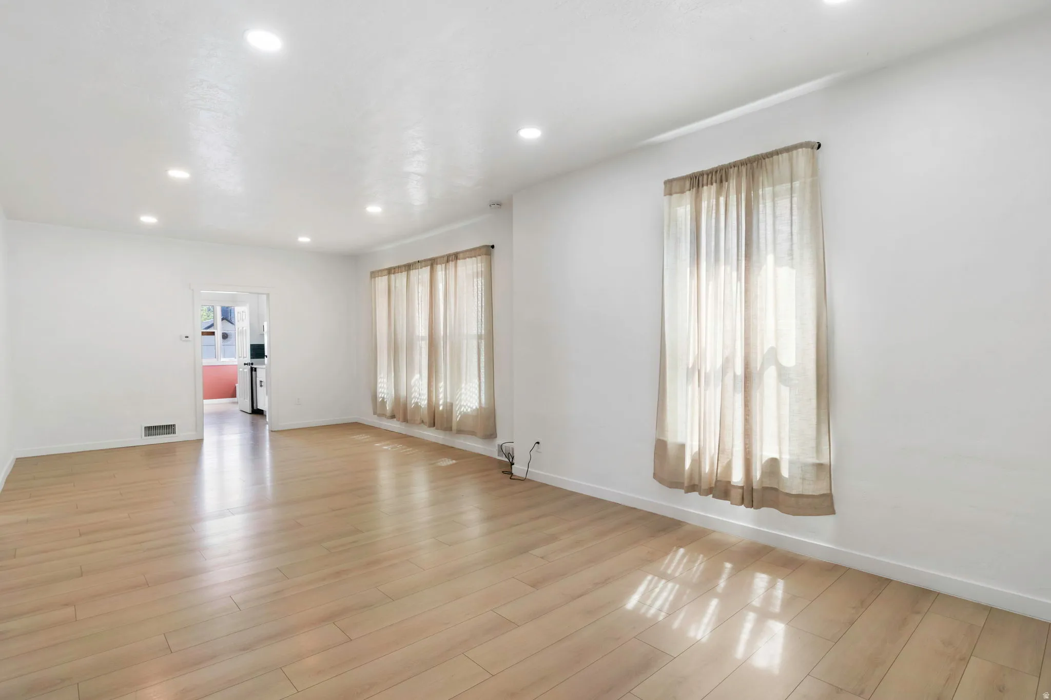 Spacious living area featuring light wood-finish flooring, recessed lighting, and multiple windows with sheer drapery