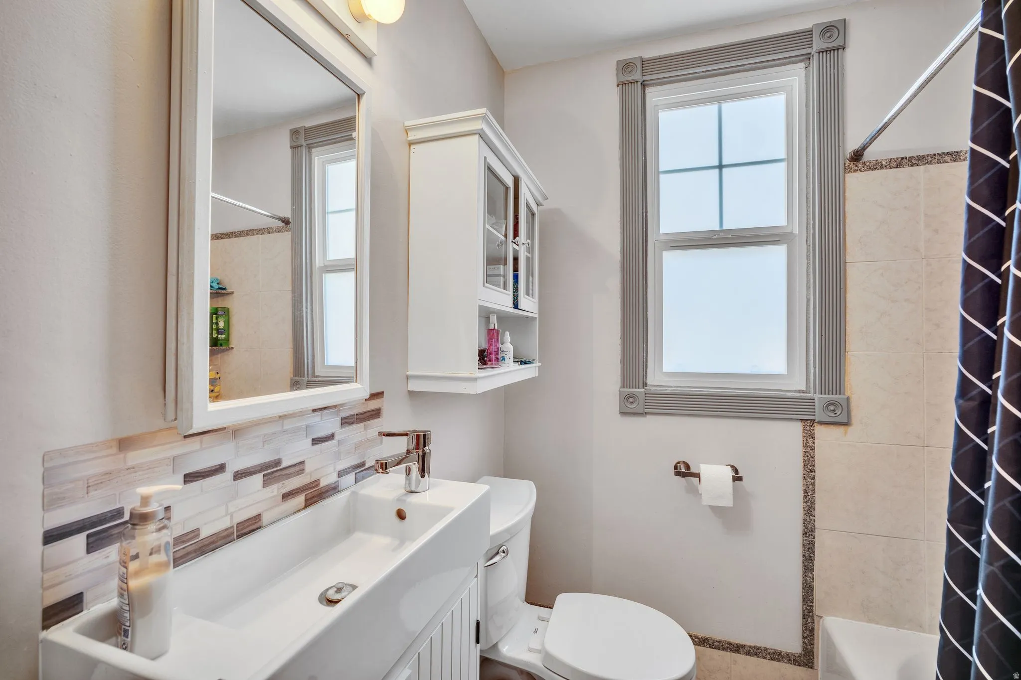 1 bedroom floorplan (618) bathroom featuring a wall-mounted medicine cabinet with integrated lighting, a rectangular vessel sink with a single-lever faucet, a tiled backsplash, an ornate window frame, and a tiled shower surround