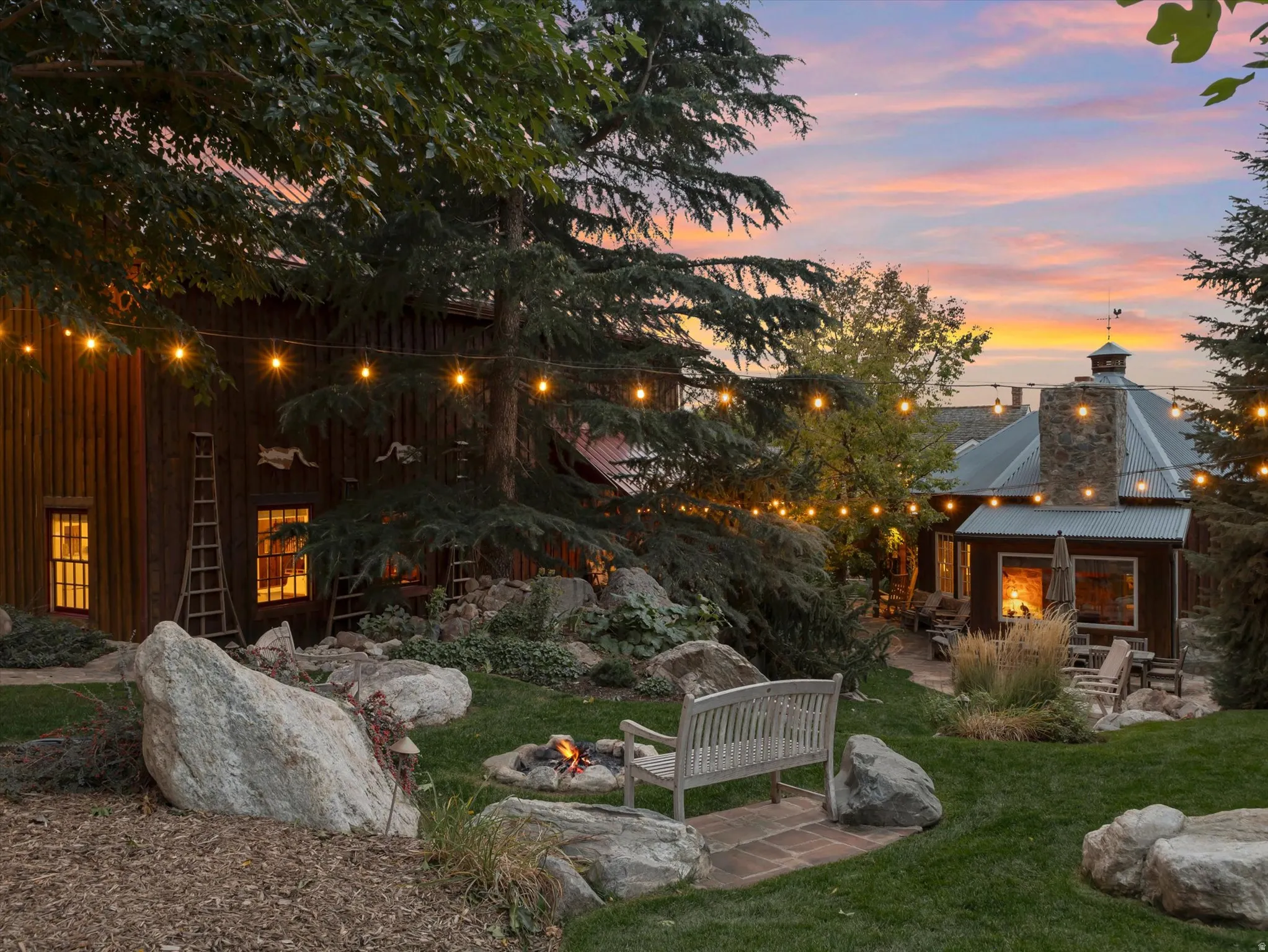 Expansive grassy area featuring a built-in fire pit, stone patio, and mature trees