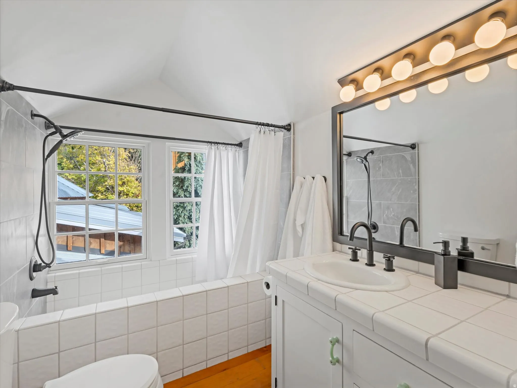 Bathroom featuring a tiled bathtub surround with dual windows, a black shower fixture, and a vaulted ceiling