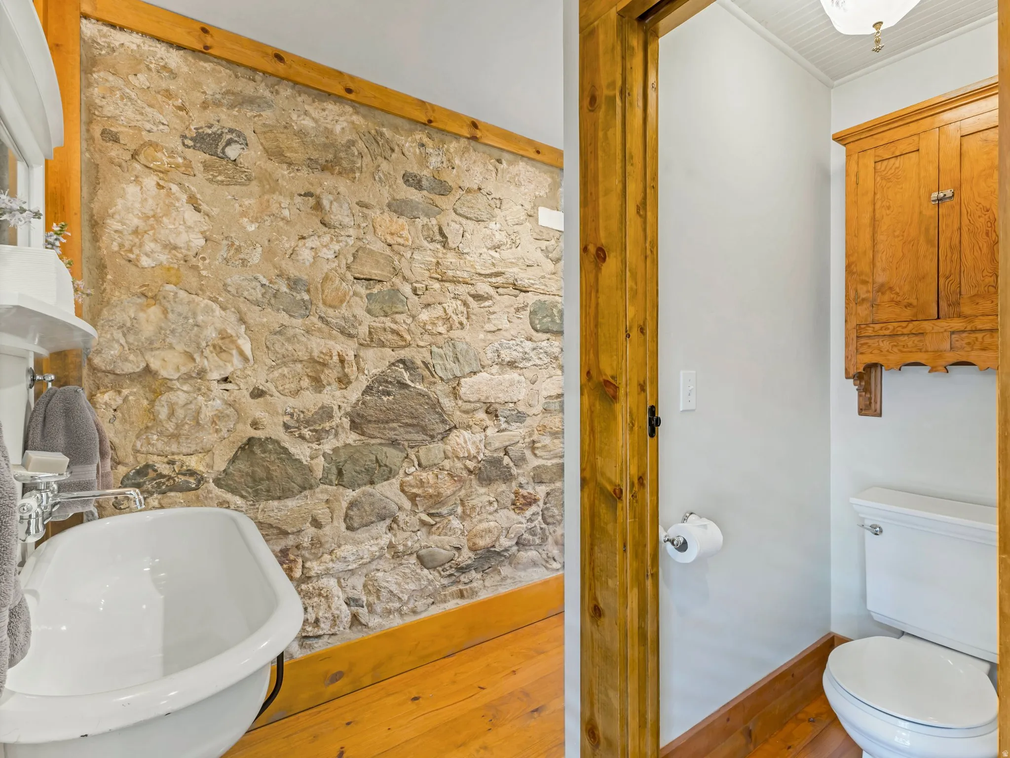 Exposed stone wall detailing a white pedestal sink with polished chrome fixtures and light wood-finish flooring