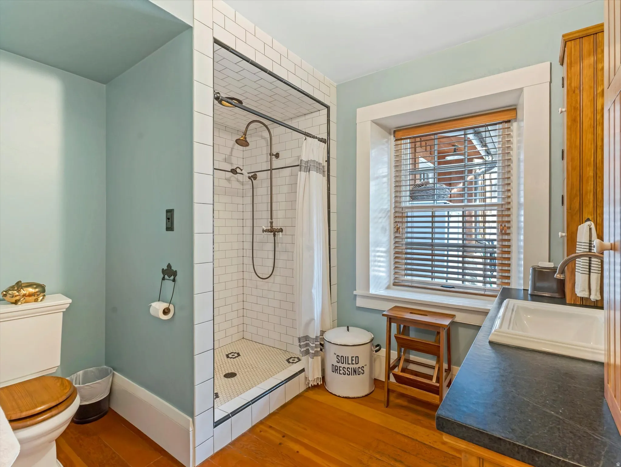 Bathroom featuring a tiled shower with a dual shower head system, wood-finish flooring, a window with natural light, a dark countertop with an integrated sink, and an exposed wood vanity