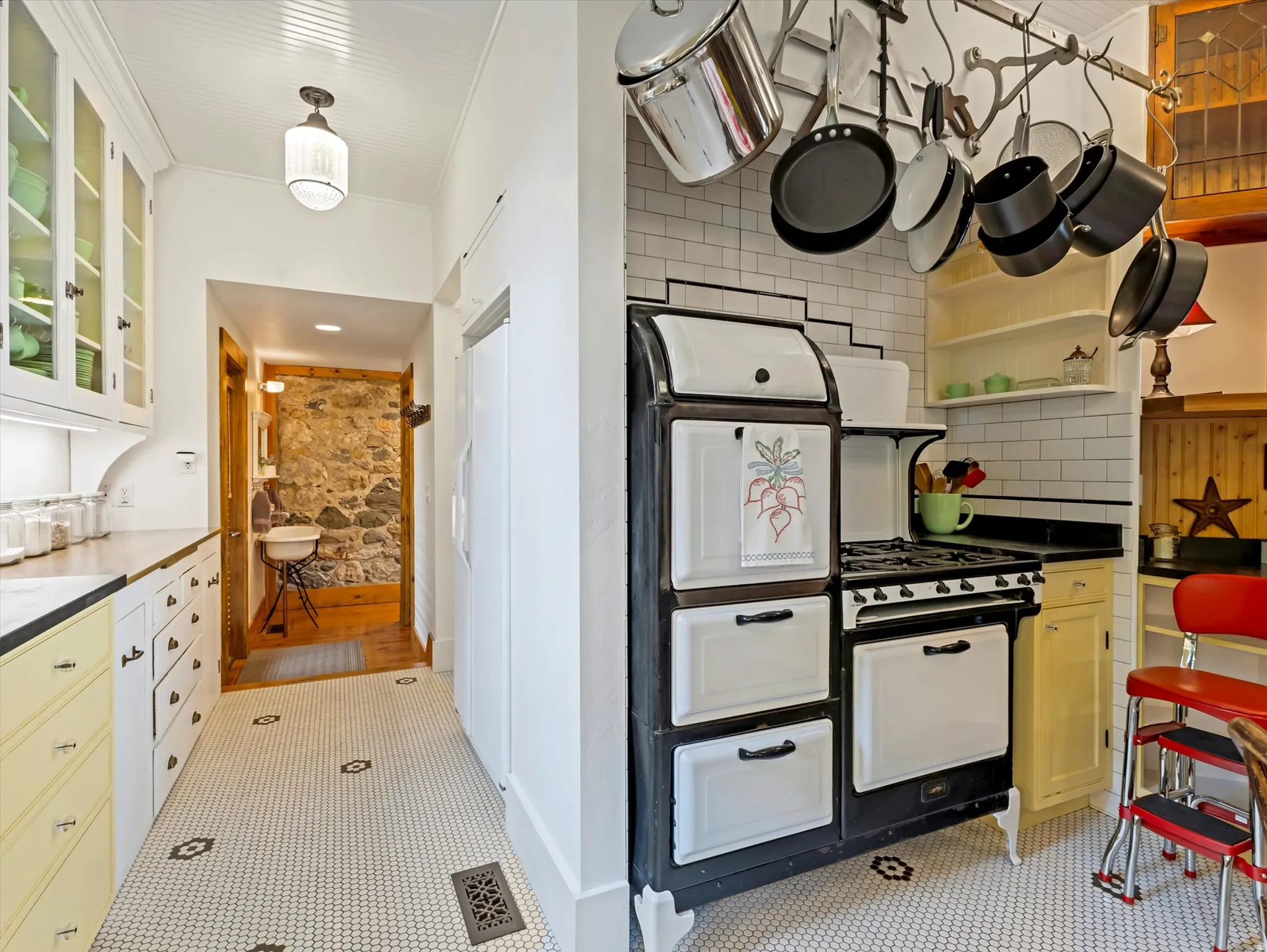 Kitchen featuring white hexagon tile flooring, a vintage-style range with white and black enamel finish, a pot rack, white subway tile backsplash, and a stone accent wall in the hallway