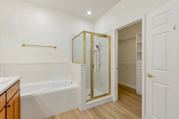 Bathroom featuring a built-in tub, a glass-enclosed shower with gold-tone framing, light wood-finish flooring, and a vanity with wood cabinetry