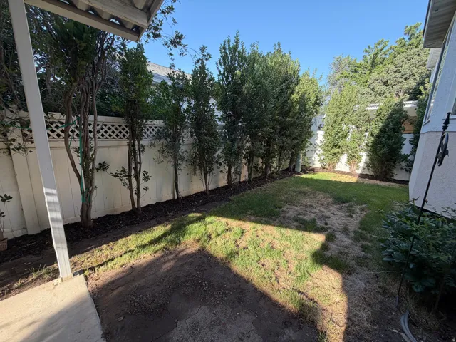 Enclosed backyard featuring a partially covered patio area, a perimeter of mature trees, a white lattice-top fence, and a grass lawn