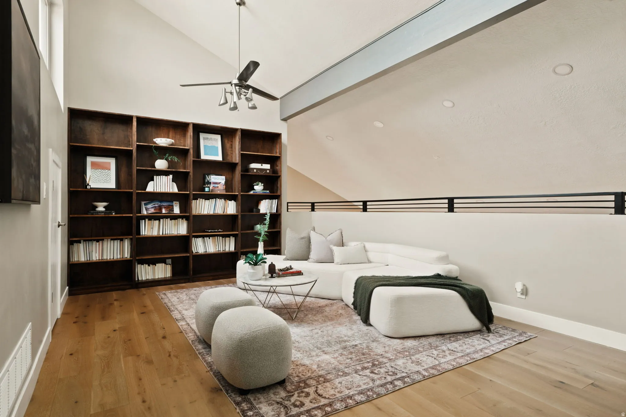 Living area featuring soaring vaulted ceilings, extensive built-in dark wood shelving, wood-finish flooring, a ceiling fan with integrated lighting, and a black metal railing overlooking the space