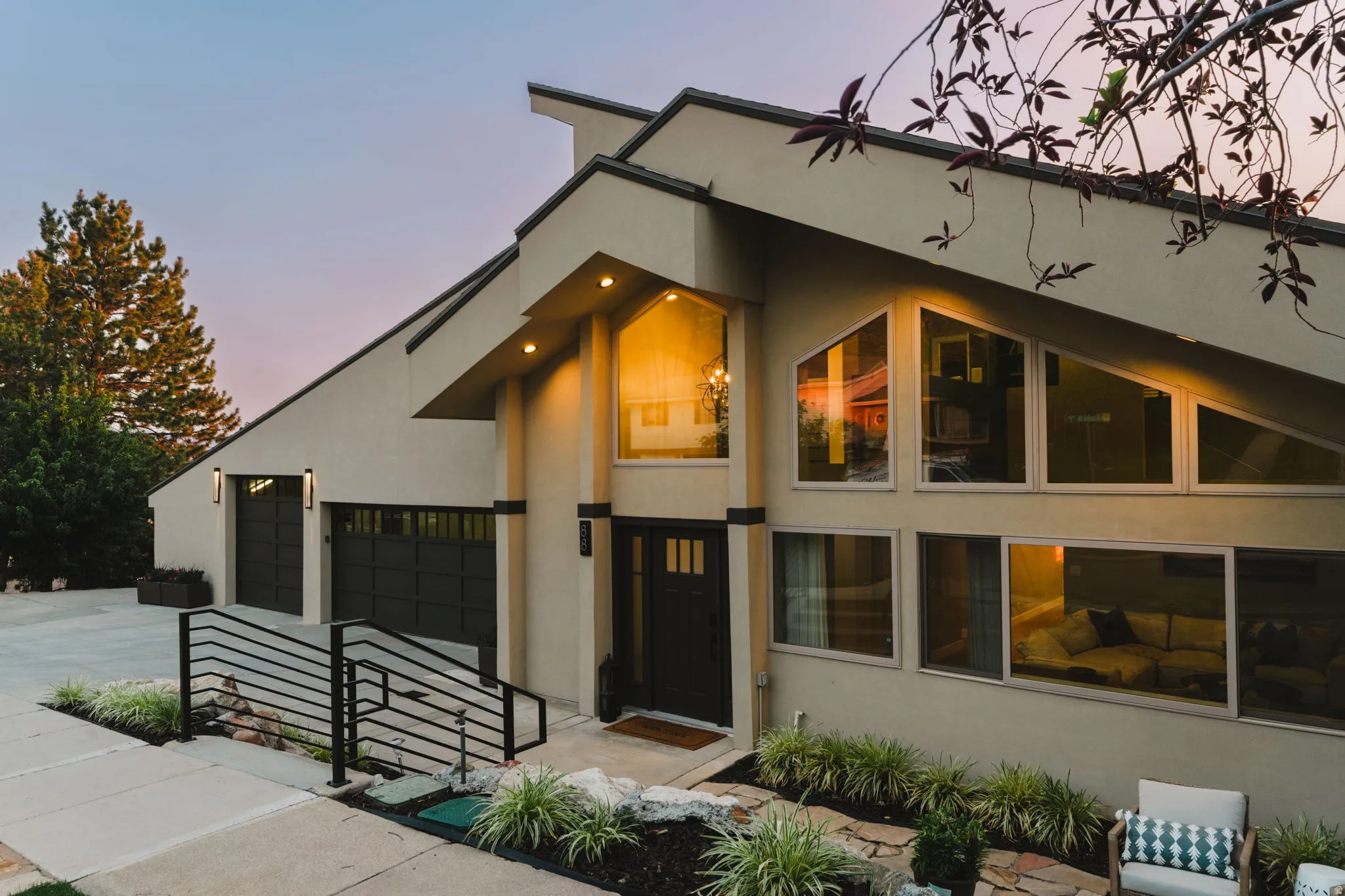 Contemporary stucco exterior featuring a prominent two-car garage with black paneled doors, multiple angular windows, a dark framed entry door, and a modern black metal handrail