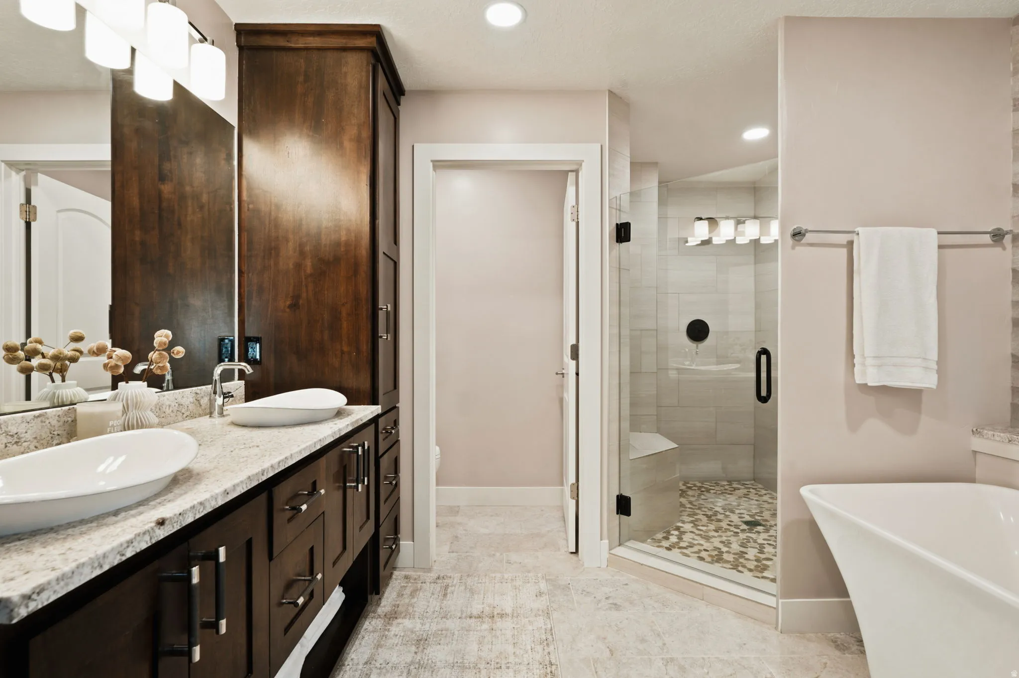 Bathroom featuring a freestanding tub, glass-enclosed shower with pebble flooring and built-in bench, dual vanity with vessel sinks, granite-finish countertop, and dark wood-finish cabinetry
