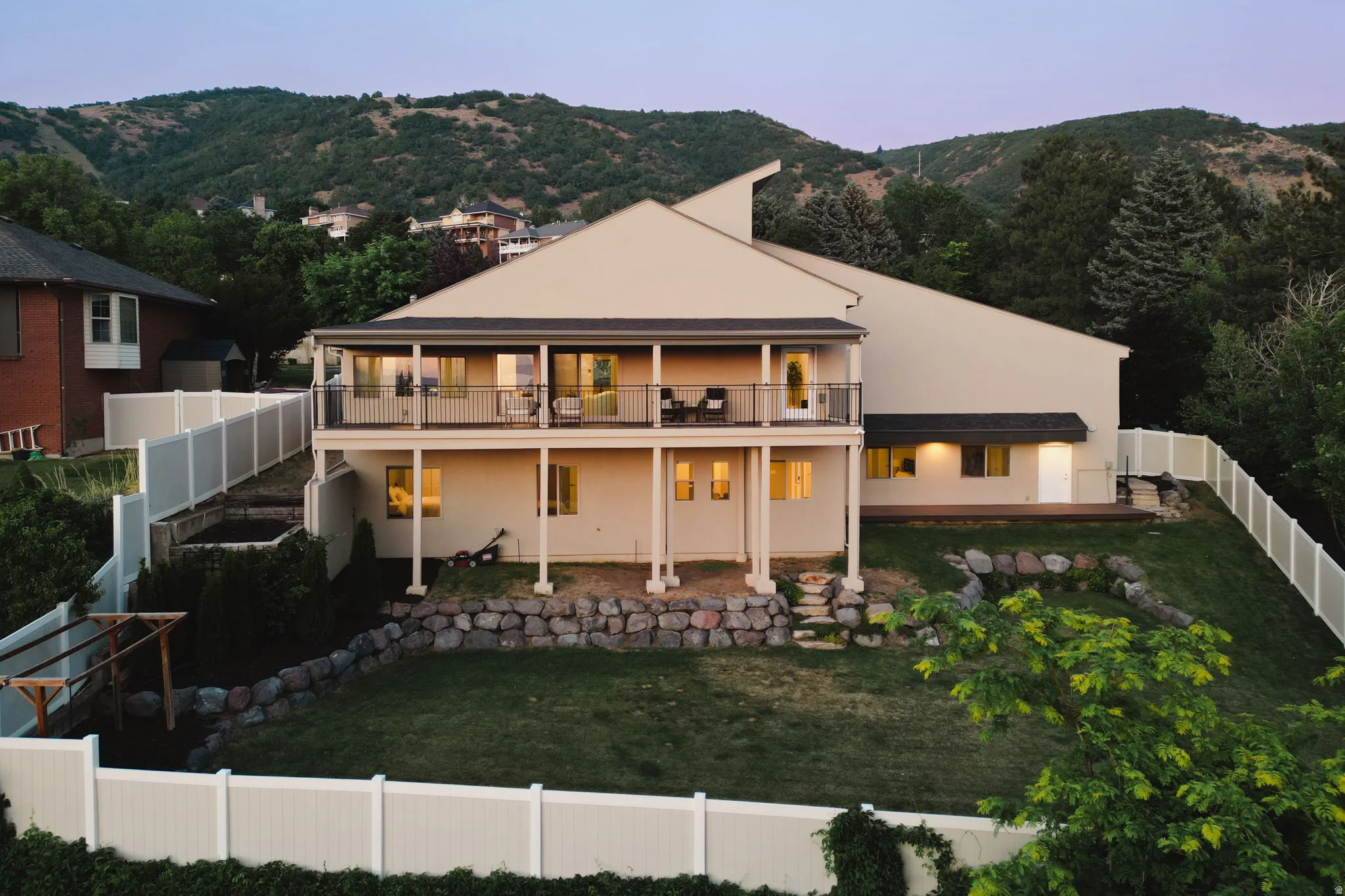 Rear exterior featuring a full-width upper-level balcony with metal railings, a lower-level walk-out patio, a sloped lawn, and a privacy fence