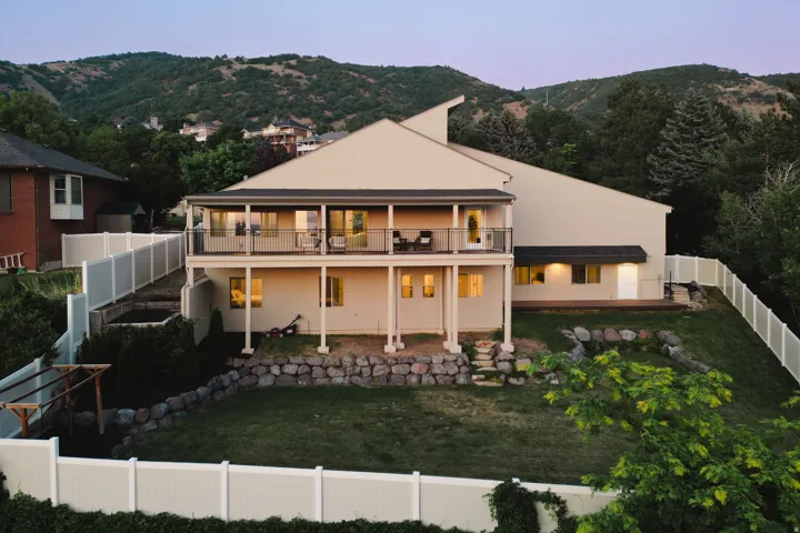 Rear exterior featuring a full-width upper-level balcony with metal railings, a lower-level walk-out patio, a sloped lawn, and a privacy fence