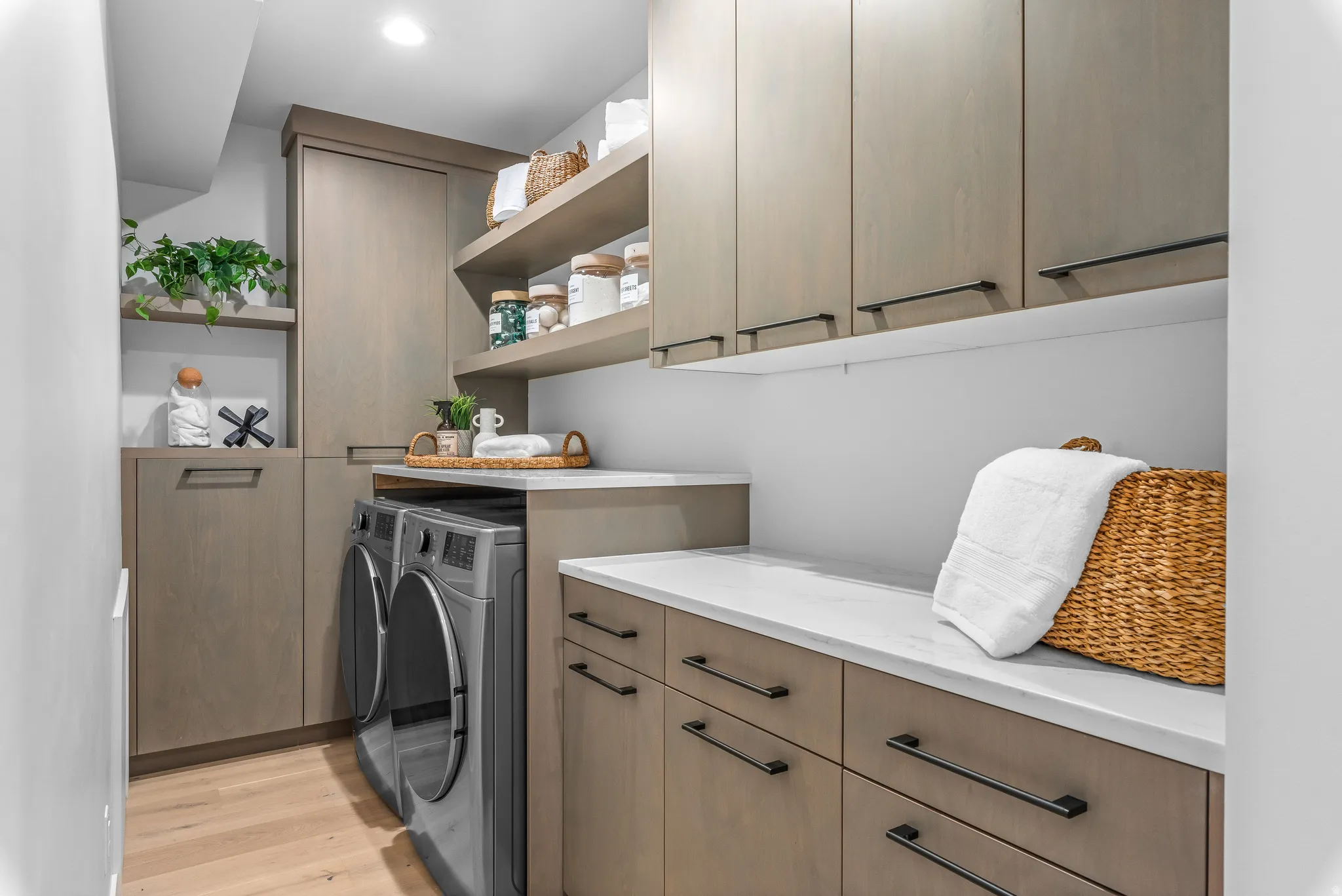Dedicated laundry space featuring stacked appliances, extensive cabinetry with dark hardware, open shelving, white countertops, and wood-finish flooring