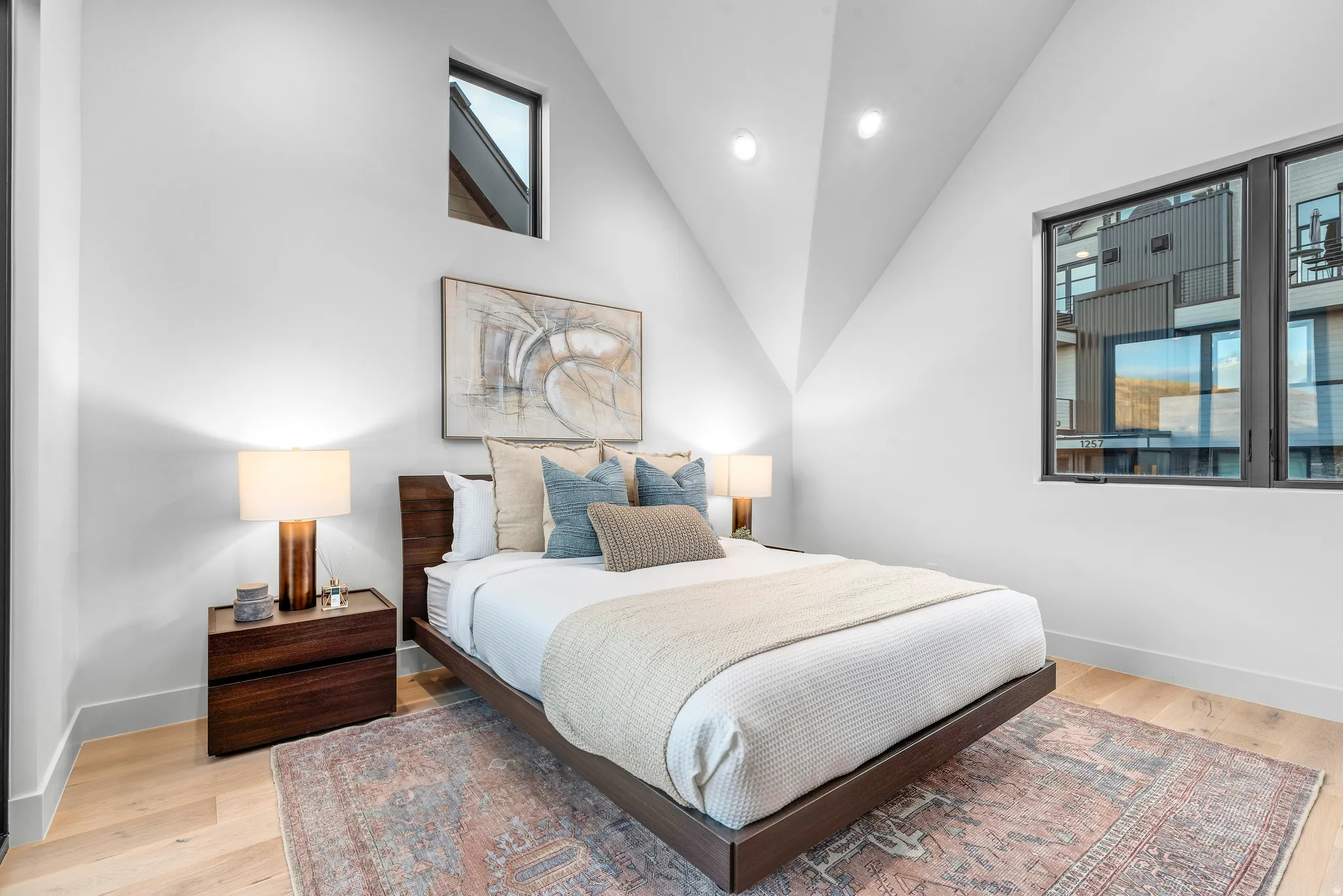 Vaulted ceiling bedroom featuring light wood-finish flooring, a large black-framed window, a small upper accent window, and recessed lighting