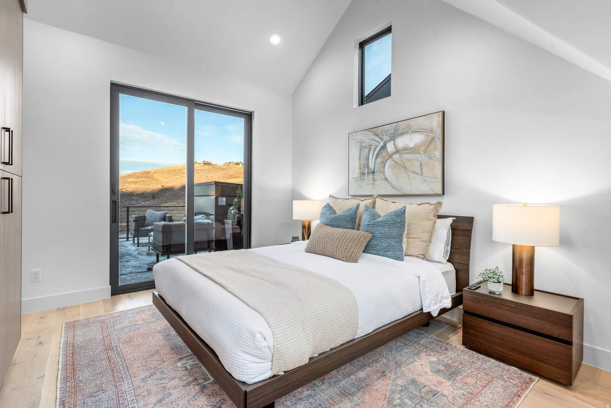 Vaulted ceiling bedroom featuring a private balcony through sliding glass doors, a high architectural window, light wood-finish flooring, and built-in closet cabinetry with dark hardware
