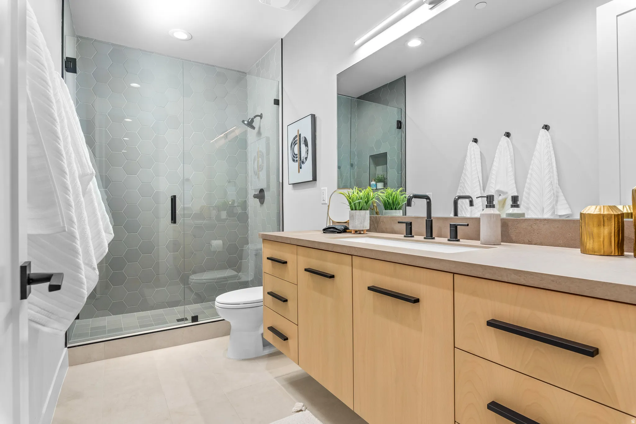 Contemporary bathroom featuring a glass-enclosed shower with hexagon tile surround, a light wood-finish vanity with ample storage, a solid surface countertop, and black fixtures