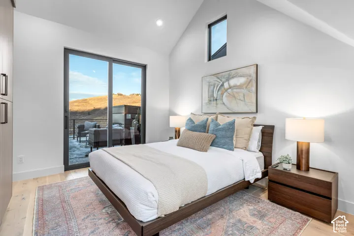Vaulted ceiling bedroom featuring a private balcony through sliding glass doors, a high architectural window, light wood-finish flooring, and built-in closet cabinetry with dark hardware
