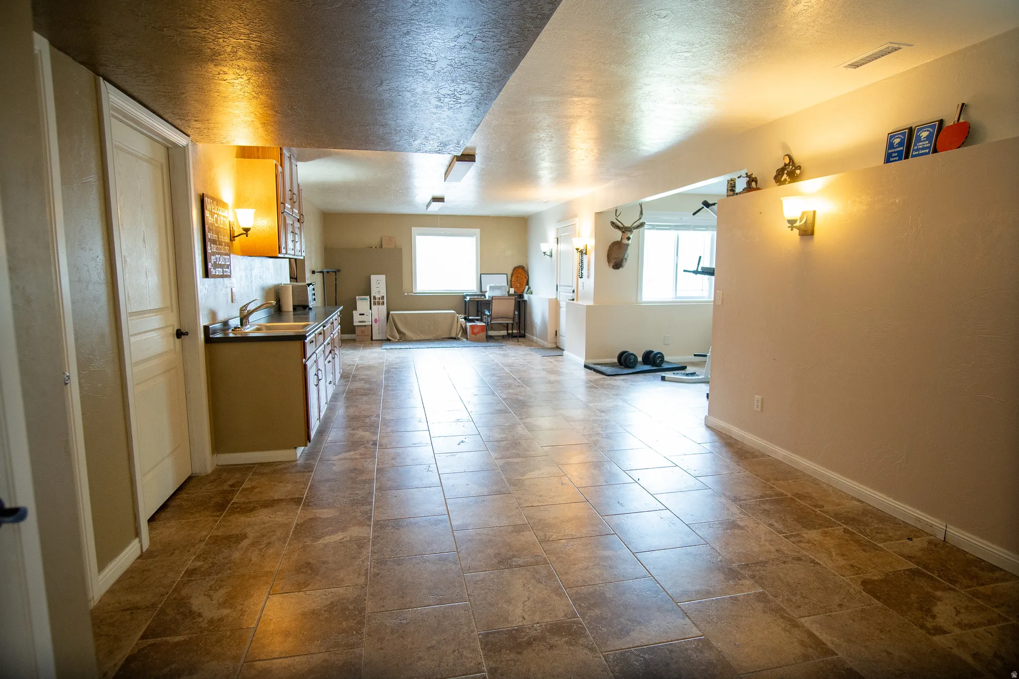 Spacious finished lower level featuring large format tile flooring, a kitchenette with dark countertops and upper/lower cabinetry, and multiple windows providing natural light