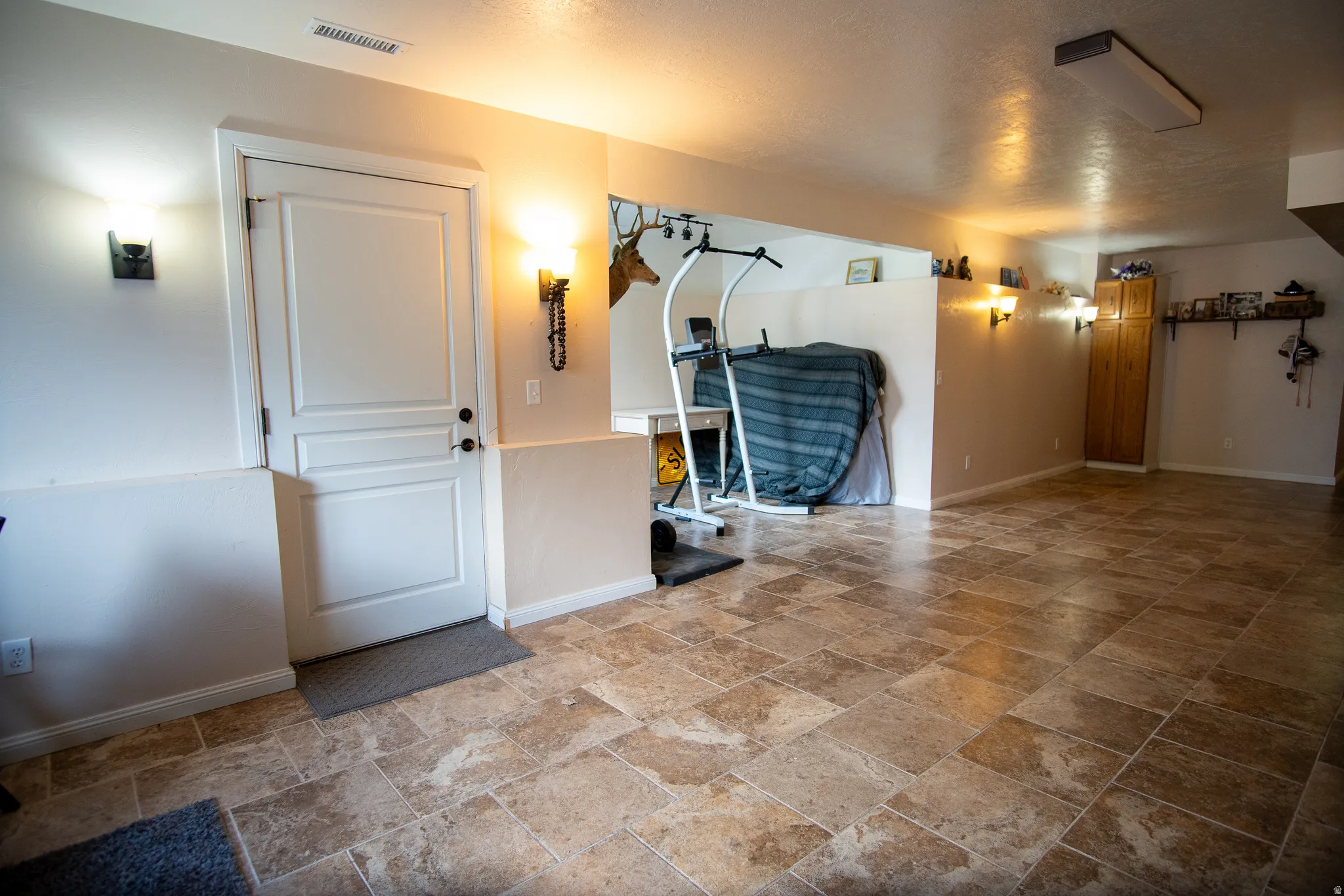 Spacious room featuring large format tile flooring, a white paneled door, and multiple wall sconces providing illumination