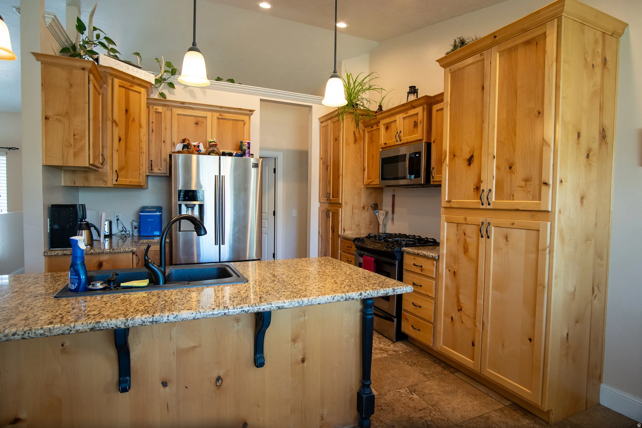 Kitchen featuring light wood cabinetry, a central island with a built-in sink, granite countertops, stainless steel appliances, and pendant lighting