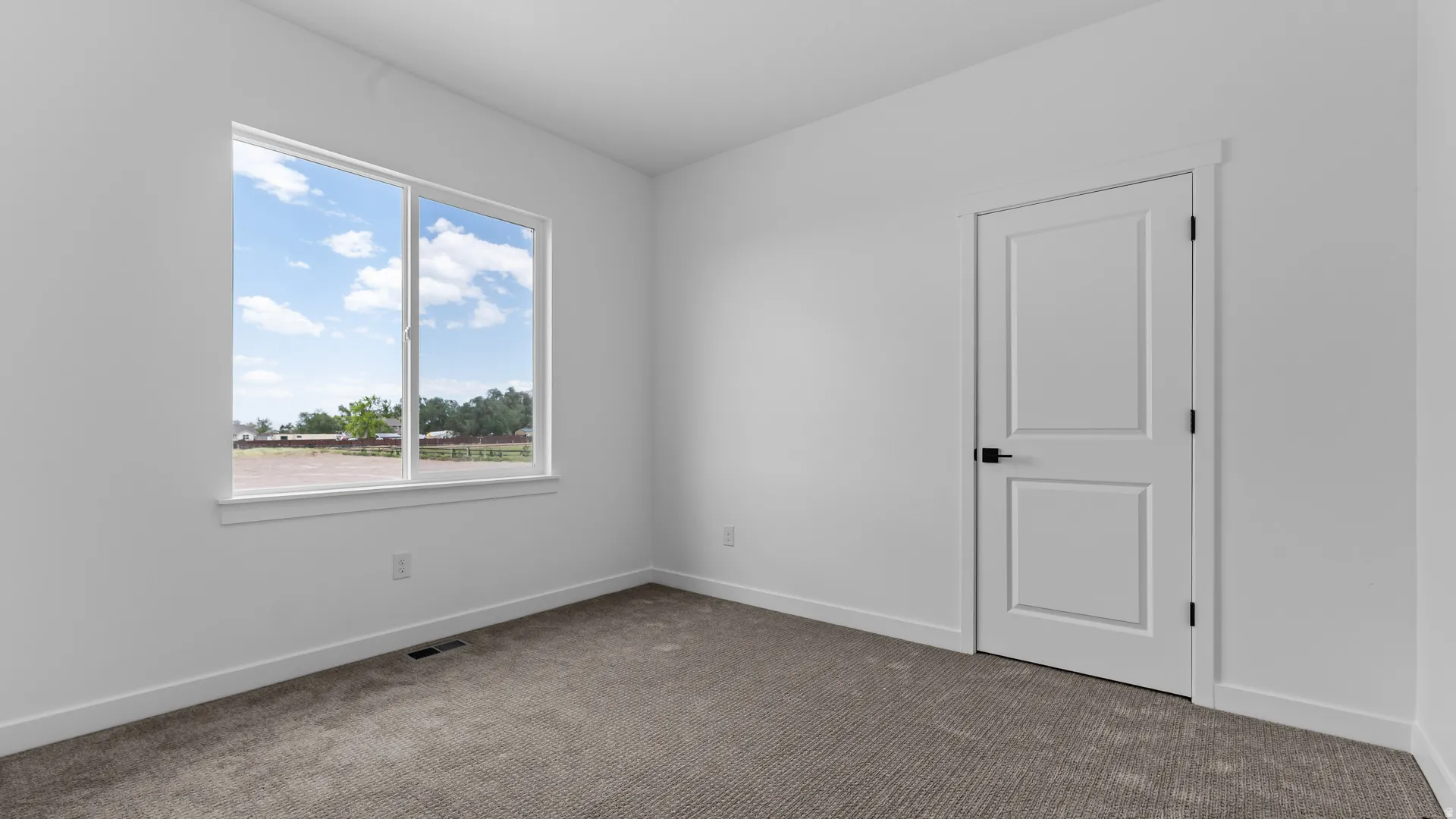 Bright room featuring a large window with white trim, fresh white walls, a two-panel interior door with matte black hardware, and light gray carpet flooring