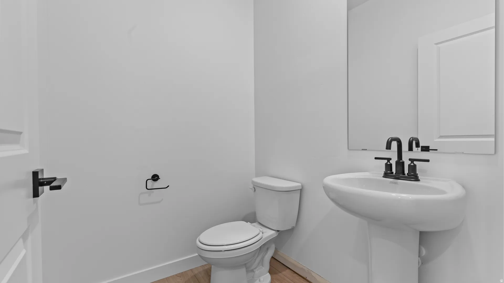 Powder room featuring a pedestal sink with matte black faucet, wall-mounted mirror, and wood-finish flooring