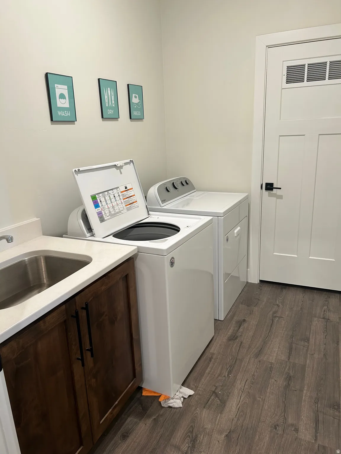 Dedicated laundry area featuring wood-finish flooring, a stainless steel utility sink, white countertop, and dark wood cabinetry with matte black hardware