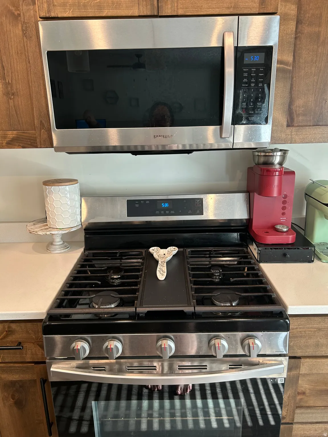 Stainless steel gas range with a built-in griddle and coordinating stainless steel microwave, set against light wood cabinetry and light countertops