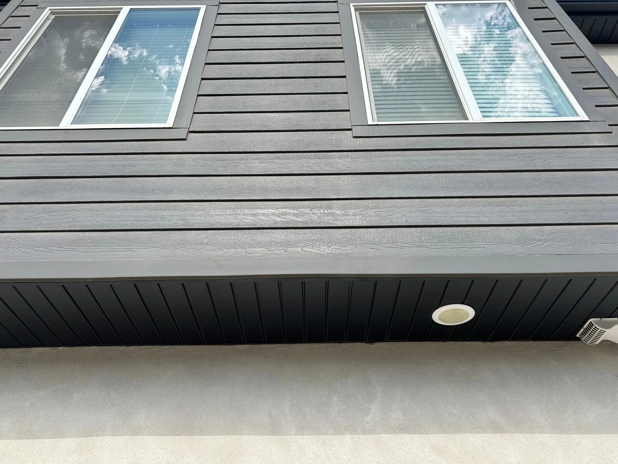 Dark horizontal siding with white-framed windows, a black soffit featuring a recessed light, and a textured foundation