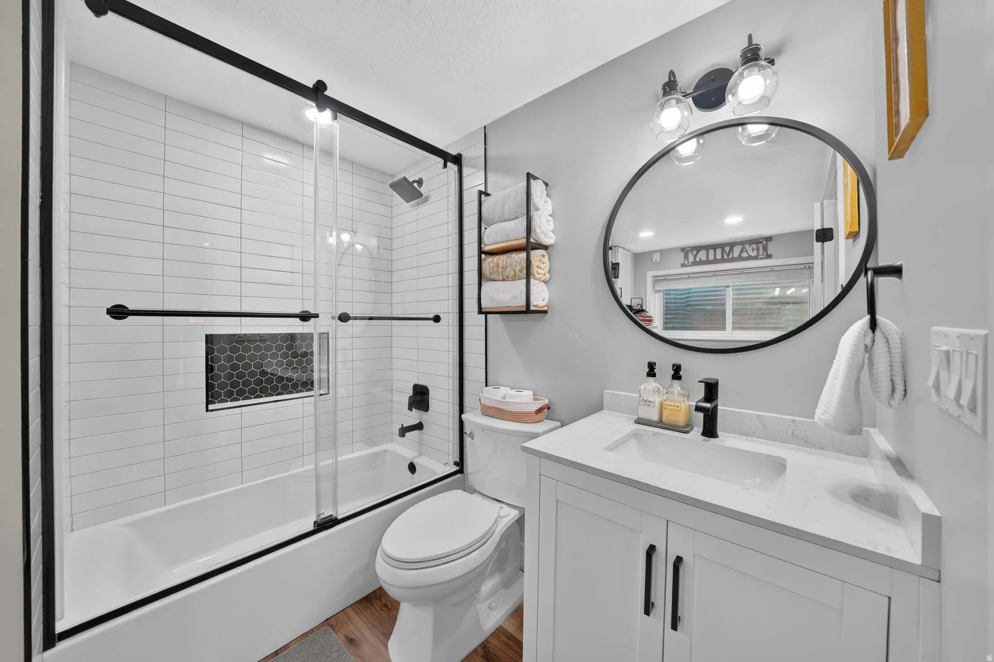Full bathroom featuring a white subway tile shower surround with a built-in hexagonal tile niche, a glass sliding shower door with black hardware, a white vanity with a single basin sink, a large circular mirror with black trim, and wood-finish flooring