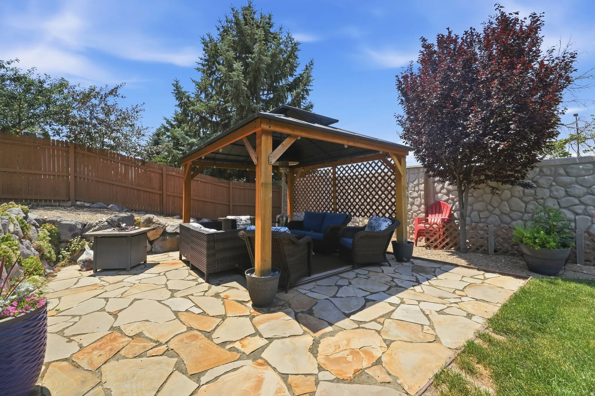 Expansive flagstone patio featuring a wooden gazebo with lattice accents, stone retaining walls, a wood privacy fence, and mature trees