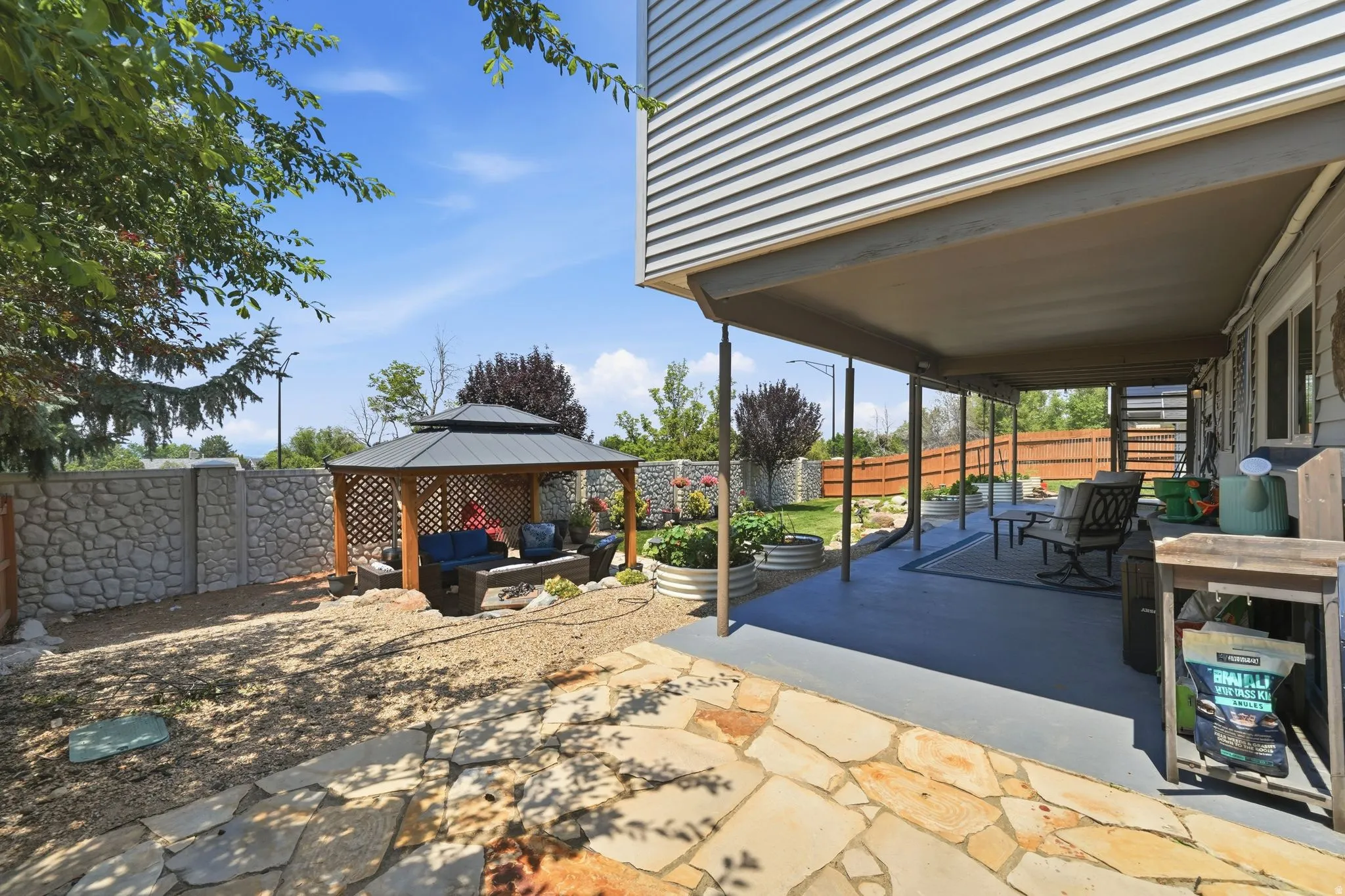 Expansive outdoor area featuring a covered patio, flagstone pathway, a gazebo with lattice detailing, stone-patterned fencing, and raised garden beds