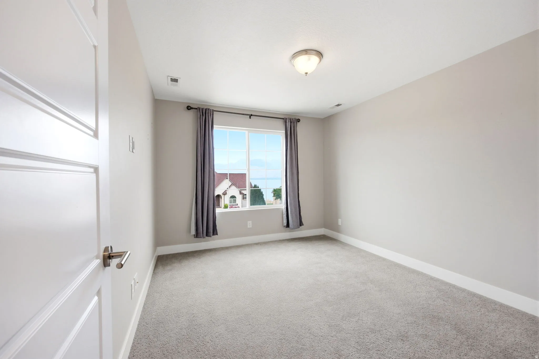 Spacious room featuring neutral paint, gray carpeting, a window with exterior scenery, a white paneled door with a brushed nickel handle, and a semi-flush mount ceiling light fixture