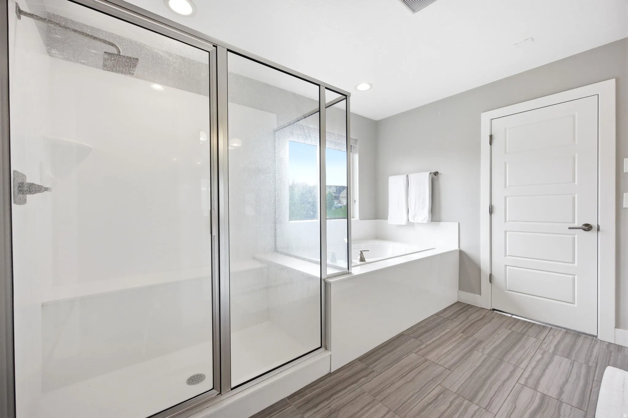 Walk-in shower with clear glass enclosure and built-in bench, soaking tub with surrounding ledge, window, and wood-finish flooring