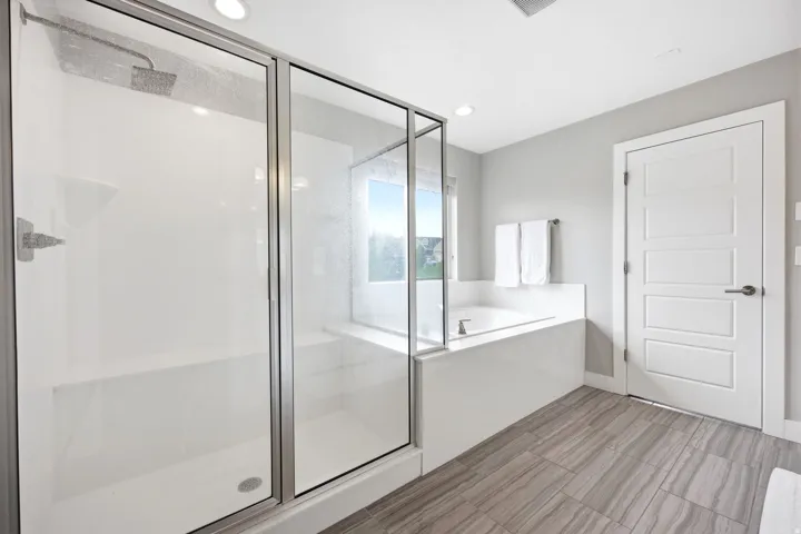 Walk-in shower with clear glass enclosure and built-in bench, soaking tub with surrounding ledge, window, and wood-finish flooring