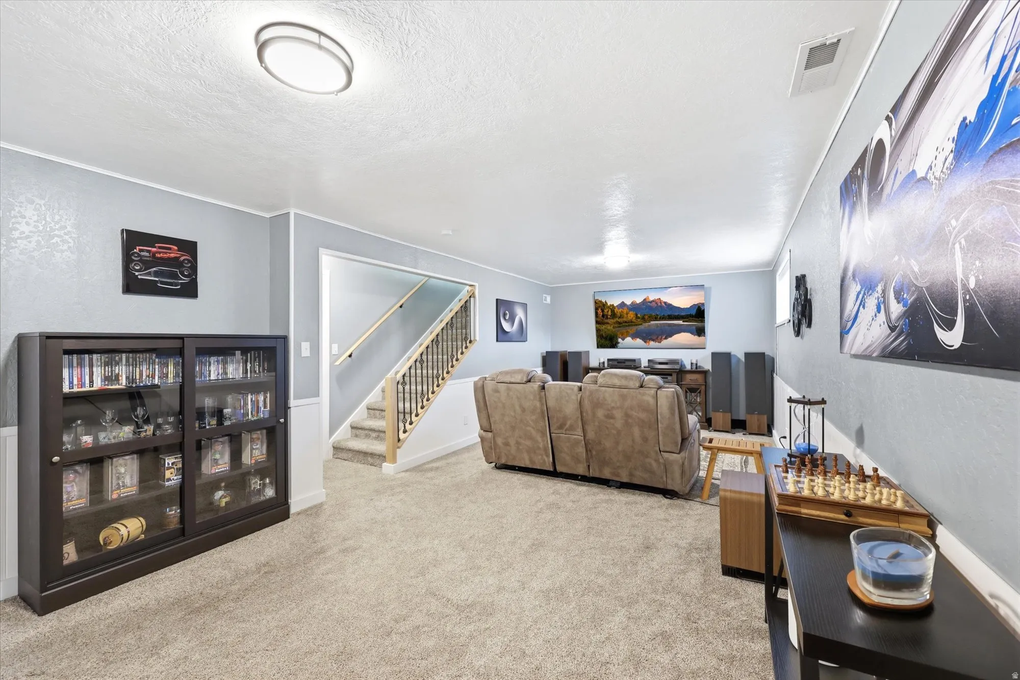 Carpeted lower-level room featuring a built-in display cabinet with sliding glass doors, a staircase with wood handrails, a wall-mounted television, and flush-mount lighting fixtures
