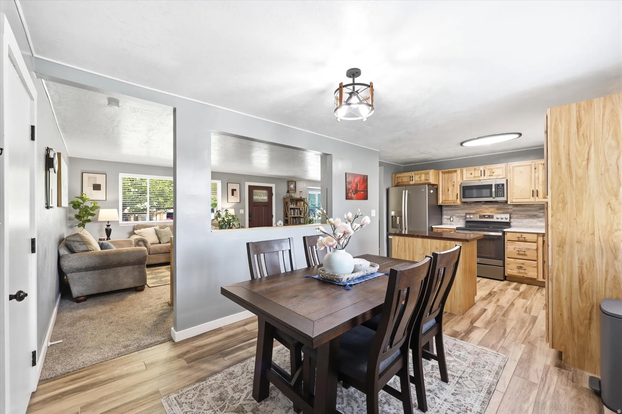 Open-concept kitchen and dining area featuring wood-finish flooring, light wood cabinetry, stainless steel appliances, and a pass-through window to an additional living space
