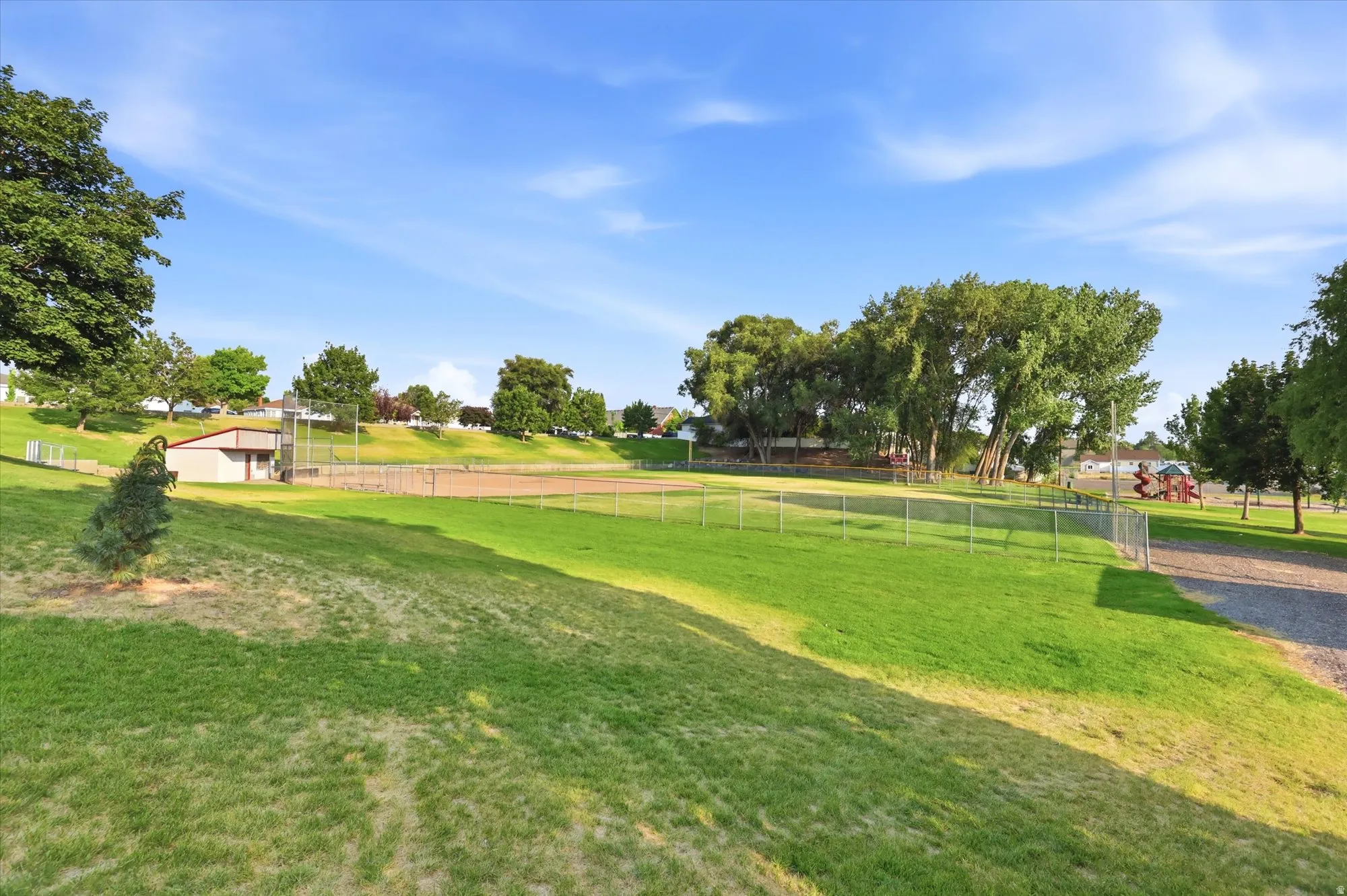 Expansive green space featuring a baseball diamond, playground equipment, mature trees, and a utility building