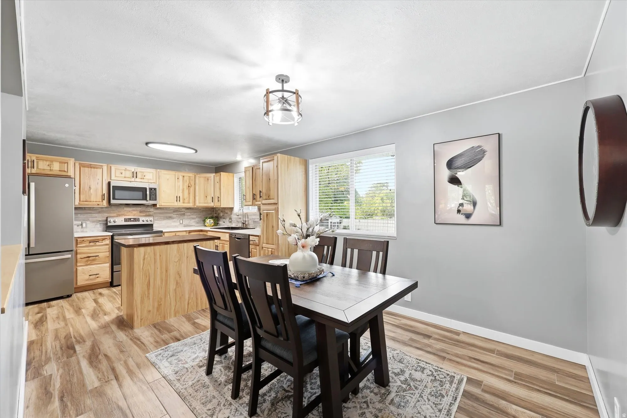 Open-concept kitchen and dining area featuring wood-finish flooring, light wood cabinetry, stainless steel appliances, a tiled backsplash, and a central kitchen island with a dark countertop