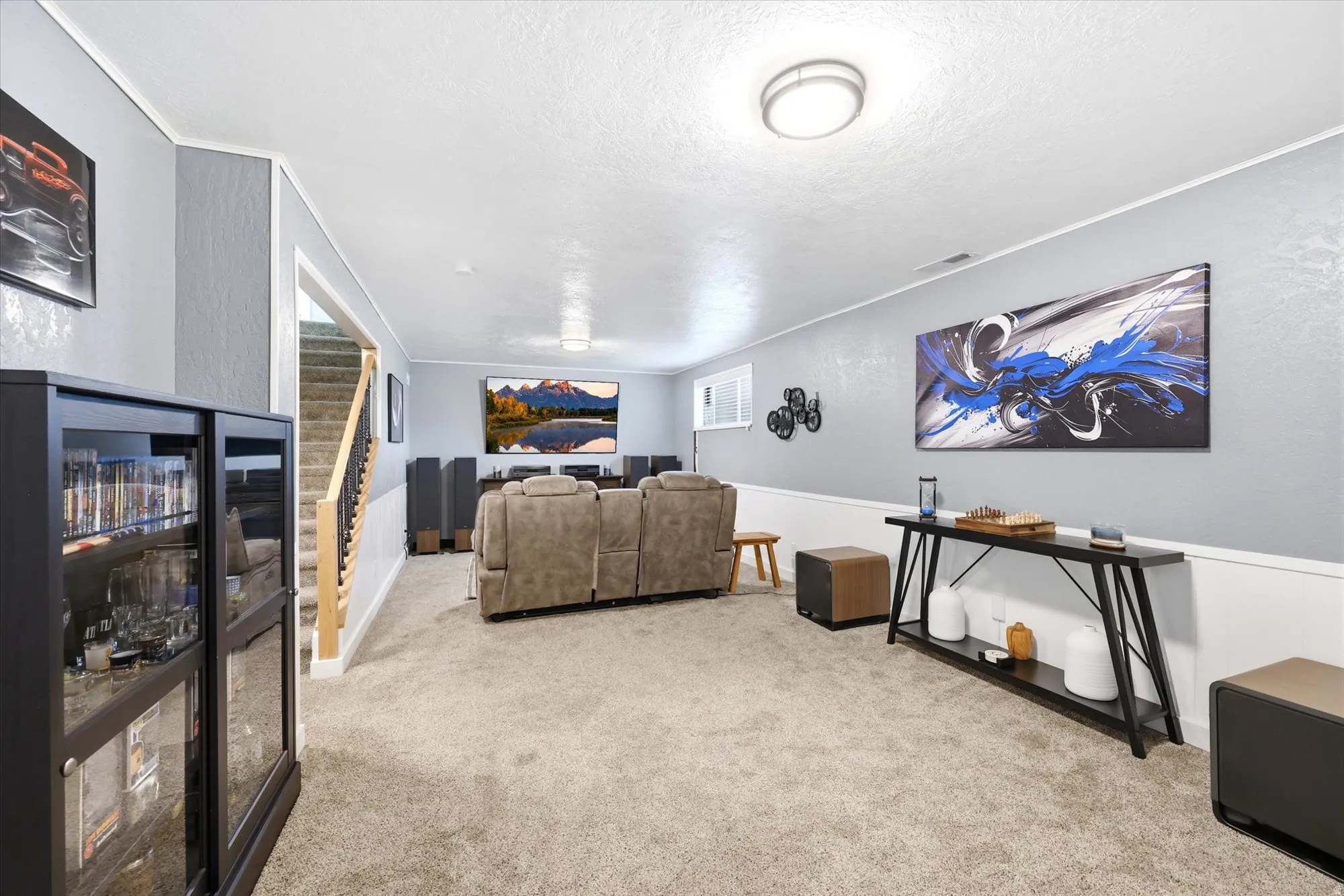 Carpeted entertainment room featuring a large wall-mounted screen, a built-in display cabinet with glass doors, and a staircase with natural wood railings
