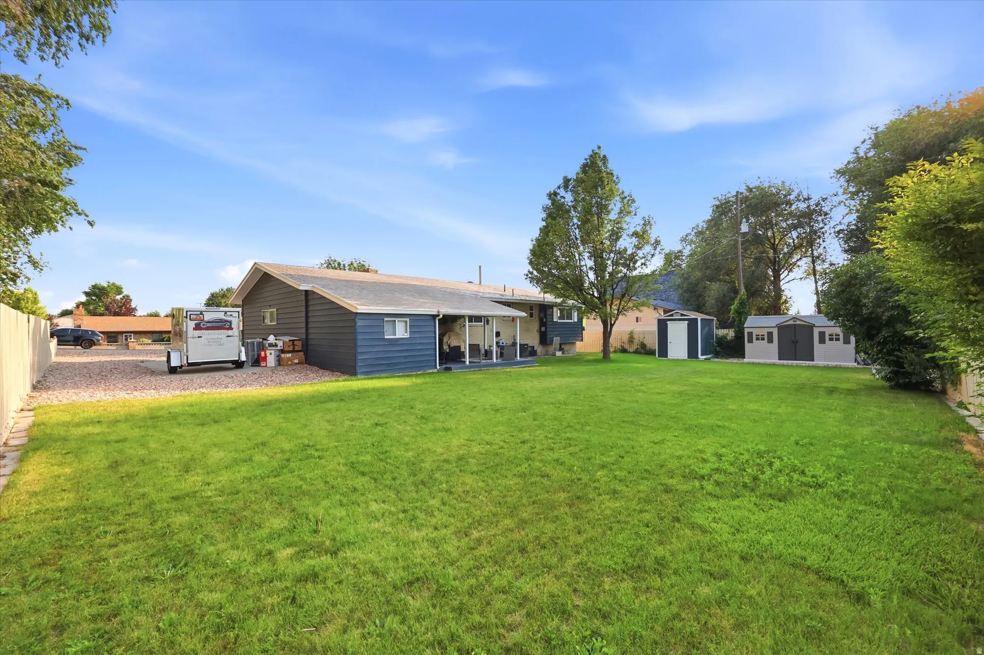 Expansive green lawn with mature perimeter trees, two storage sheds, and a gravel parking area