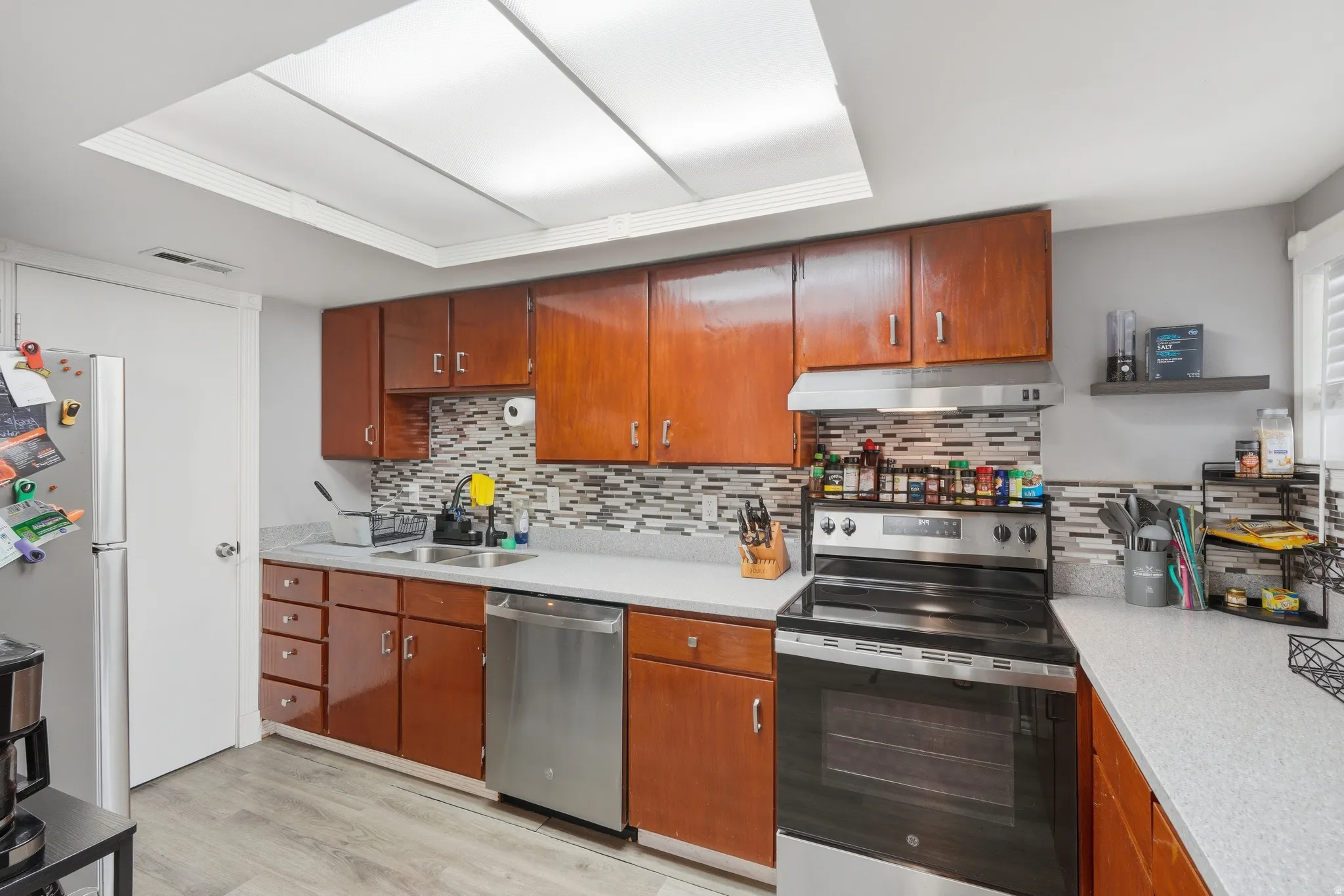 Kitchen featuring wood-finish cabinetry, stainless steel appliances, a modern tile backsplash, light-toned countertops, and wood-finish flooring