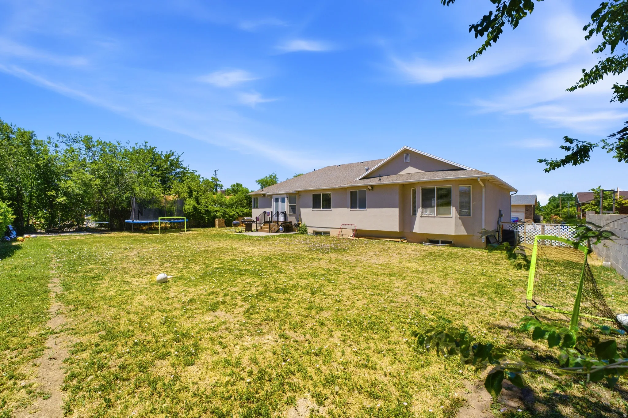 Spacious backyard featuring a large grass area, mature perimeter trees, and a beige exterior home with multiple windows
