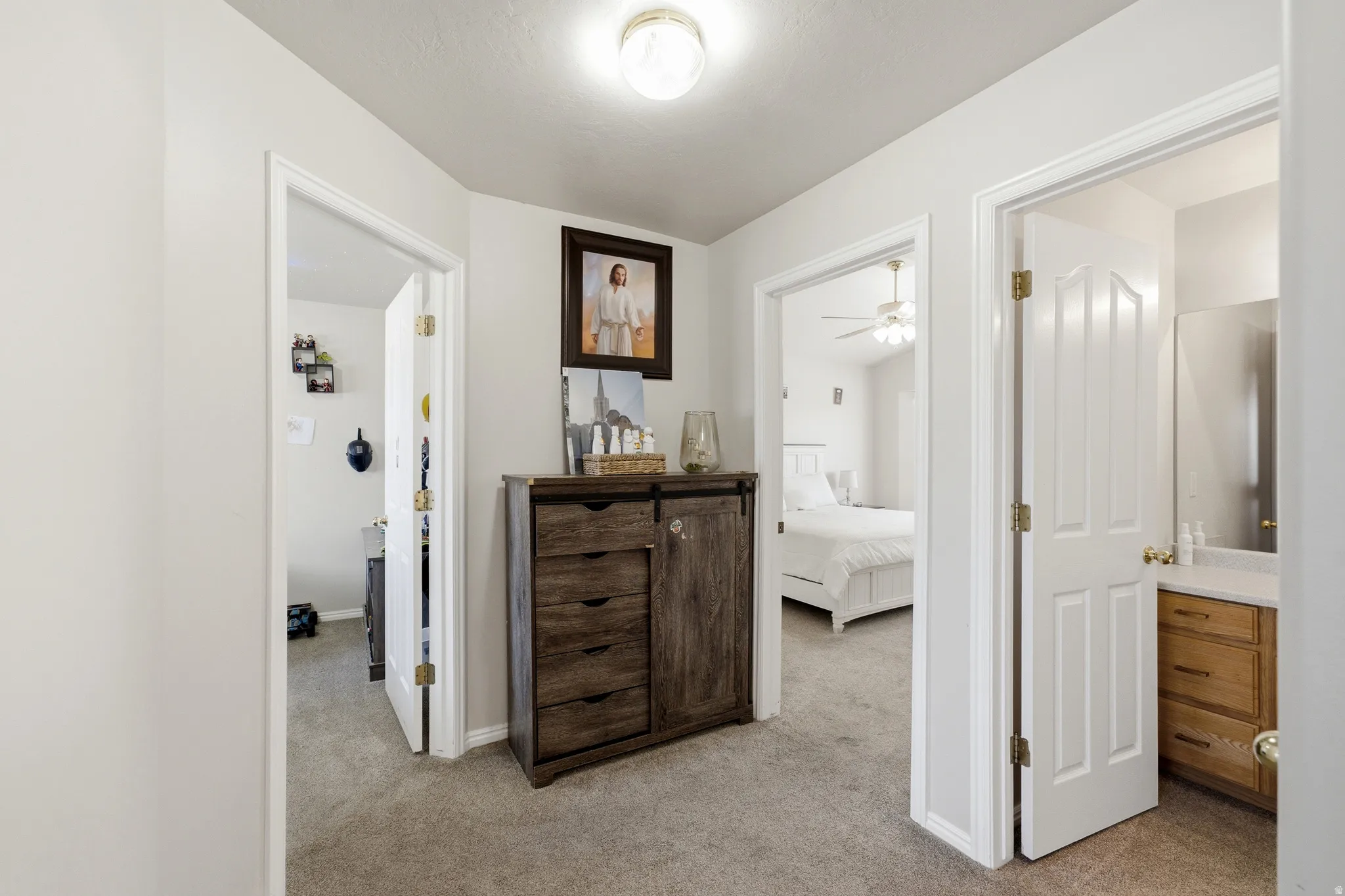 Carpeted hallway with white walls and trim, providing access to multiple rooms including a bedroom with a ceiling fan and a bathroom with a wood-finish vanity and mirror