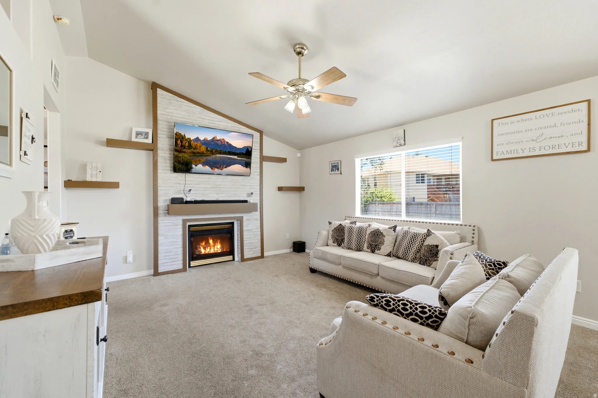 Living area featuring a vaulted ceiling with a ceiling fan, a fireplace with a white shiplap-style surround and wood-finish mantel, and light-colored carpet flooring