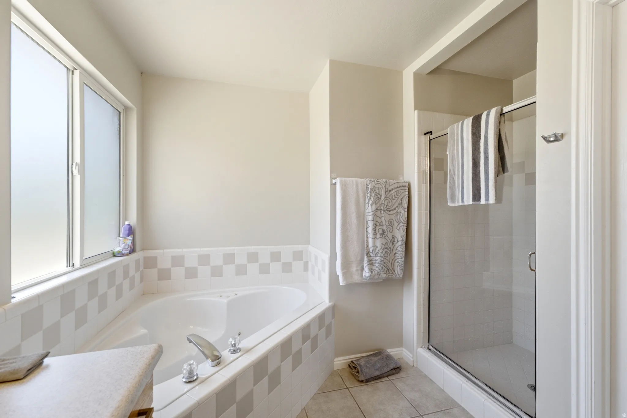 Bathroom featuring a built-in corner soaking tub with tiled surround, a separate glass-enclosed shower, a large frosted window, and neutral wall paint